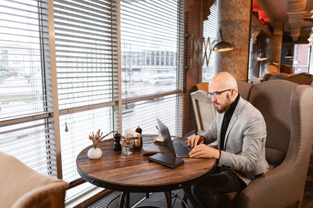 在眼镜上留着胡子的年轻人的肖像, 望着显示器的笔记本电脑。成功人士, 商人在舒适的咖啡馆。在上网本工作中阅读文本