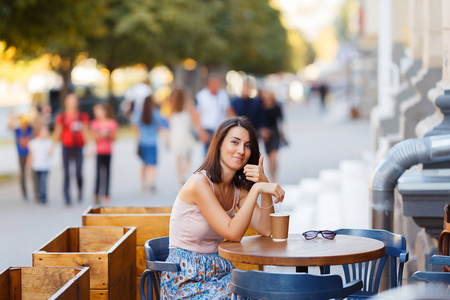 一位美丽的微笑快乐的女士的肖像，她坐在咖啡馆的夏天露台上喝着热咖啡。 一个年轻的女孩坐在餐厅的桌子上喝咖啡。