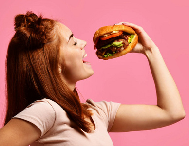 一个饥饿的年轻妇女吃汉堡的近距离画像查出在粉红色的背景