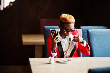 时尚非裔美国人模特穿着红色西装，头发和太阳镜，坐在咖啡馆里喝咖啡，用手机说话。