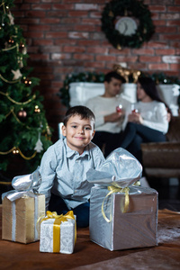 一个小男孩带着礼物盒在地板上，背景是他的父母和圣诞树