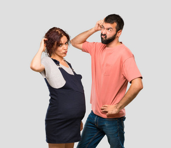 一对夫妇怀孕的妇女有疑问，并有困惑的脸，同时在孤立的灰色背景上挠头。