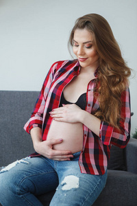 美丽的孕妇，长发穿着格子衬衫，坐在沙发上，的肚子抚摸着他的手。 幸福孕产健康生活方式的概念。