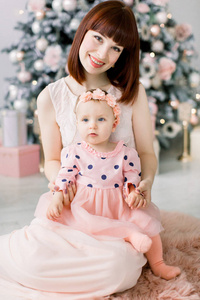 圣诞节和新年快乐的假期。 家庭母婴概念。 快乐的妈妈和她可爱的女婴穿着粉红色的裙子，玩得很开心，坐在室内圣诞树附近。