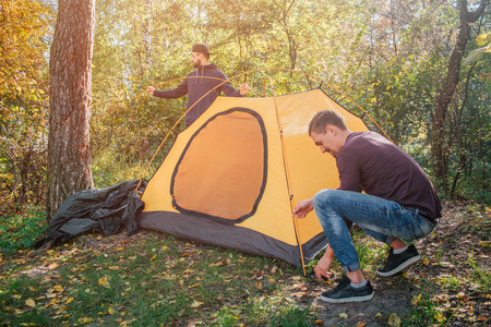 两个年轻人在森林里搭帐篷的照片。一个人用绳子工作。另一个站在帐篷后面。他们一起工作
