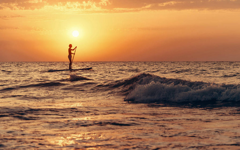 日落时，年轻人在海中的一块冲浪板上划水的剪影。