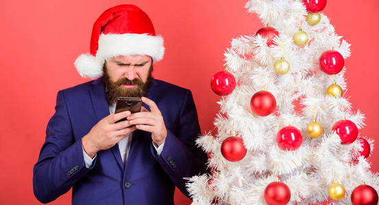 短暂的圣诞祝福。经理在网上向同事们表示祝贺。阅读圣诞问候。男人留胡子的嬉皮士穿着正式的西装和圣诞老人的帽子举行智能手机。发送圣诞