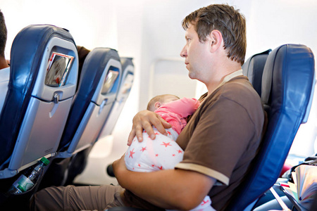 年轻人在飞行上飞机去度假期间累了父亲和他哭泣的宝贝女儿。爸爸抱着女婴在胳膊上。空中旅行与婴儿 儿童和家庭的概念