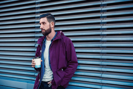 英俊的年轻人穿着紫色的冬衣喝咖啡。那个在街上喝咖啡的家伙