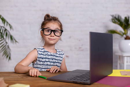可爱的小女孩坐在桌旁, 带着笔记本电脑和笔记本电脑, 戴着眼镜