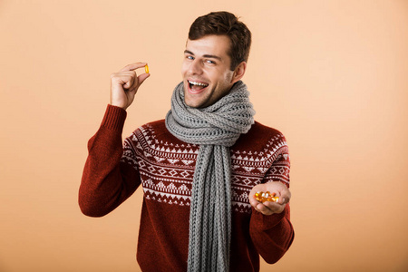 肖像一个快乐的男人穿着毛衣和围巾，被隔离在米色背景上，展示药丸胶囊