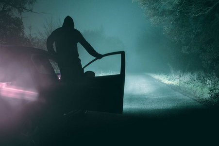 一个阴险的蒙面人站在一辆汽车上，在雾蒙蒙的冬天的夜晚剪影。 用冷蓝灰色编辑。