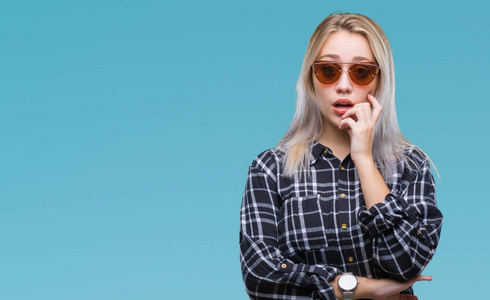 年轻的金发女人戴着太阳镜在孤立的背景上看起来压力和紧张，双手咬着嘴的指甲。 焦虑问题。