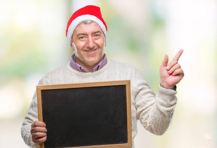 英俊的老人戴着圣诞帽，拿着黑板在孤立的背景上，非常高兴地用手和手指指向一边
