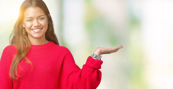 年轻漂亮的黑发女人穿着红色的冬季毛衣，在孤立的背景上微笑，欢快的呈现，用手掌指着相机。