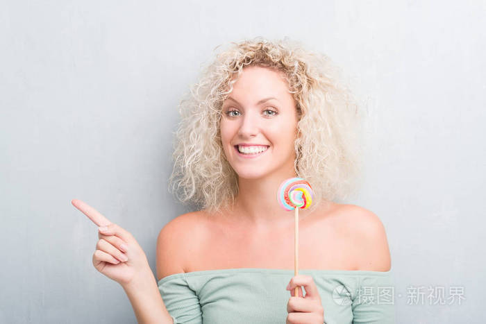 年轻的金发女人在灰色的墙壁上吃糖果棒棒糖,非常高兴地用手和手指