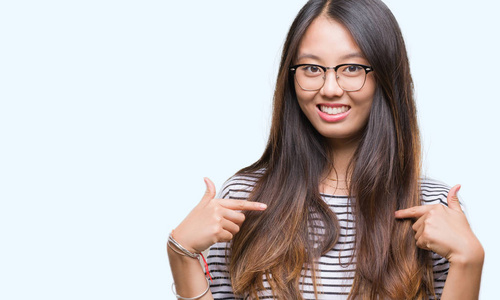 年轻的亚洲女人戴着眼镜，在孤立的背景上看着自信，脸上带着微笑，手指着自己，骄傲和快乐。