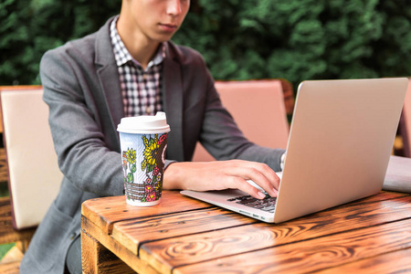 这位年轻人正坐在户外的桌子旁，手提电脑和喝咖啡