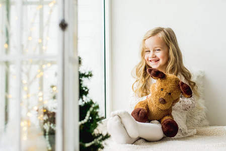 小女孩和一只毛绒鹿坐在窗户上。 一个孩子望着窗外，正在等待圣诞老人的到来