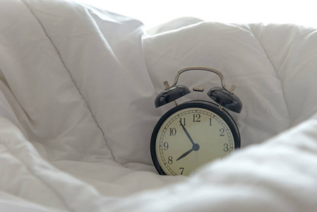 老式闹钟起床白色床单羽绒被和枕头在早晨与阳光明媚的一天。 生活方式概念