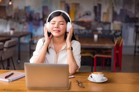 美丽的亚洲女人在咖啡馆里放松和听音乐，有笔记本电脑和咖啡杯。 人和生活方式的概念。 自由职业快乐工作场所主题。 大学和学院的主题