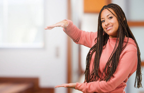 一位年轻的黑人妇女戴着辫子，用手拿着东西，展示了一种微笑和快乐的产品，提供了一个想象中的物体