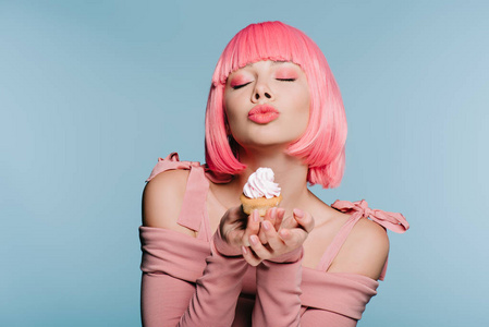 穿着粉红色假发的时髦女孩拿着甜蜜的纸杯蛋糕，吹着蓝色的吻