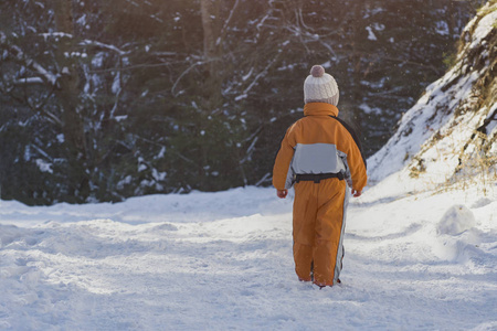 穿着橙色连衣裙的小男孩站在针叶林中覆盖着雪的道路上。 冬天阳光明媚的一天。 后景。