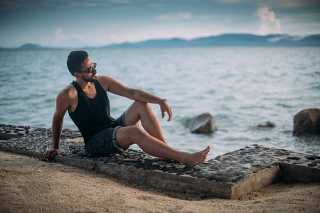 一个年轻人坐在海滩上。 戴太阳镜的帅哥在日落时看着海洋上的地平线。
