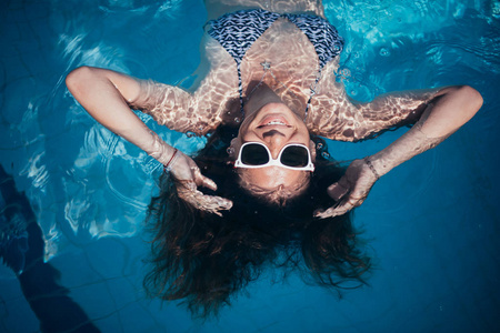一个戴太阳镜的年轻女孩在游泳池里游泳。 美丽的女人在室外游泳池的水里休息