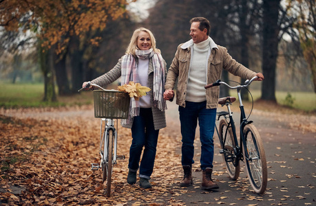 高级夫妇秋天在公园散步。 有魅力的女人和英俊的男人在一起玩自行车
