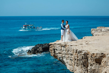 美丽的微笑，年轻的新娘和新郎在海滩上散步，亲吻，在岩石和大海附近举行有趣的婚礼。 塞浦路斯海岸婚礼