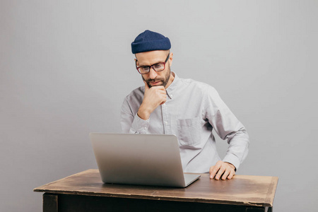 男性自由职业者在互联网上阅读新闻，专注地看笔记本电脑，看视频，坐在旧桌子上，穿着白色衬衫，头饰被隔离在灰色的墙上。 商人思考创业