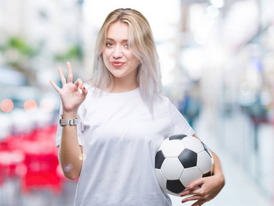年轻的金发女子在孤立的背景上手持足球球，用手指做好标志，很好的标志