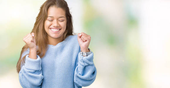 年轻漂亮的黑发女人穿着蓝色的冬季毛衣，在孤立的背景下兴奋地成功，举起双臂庆祝胜利，微笑。 赢家概念。