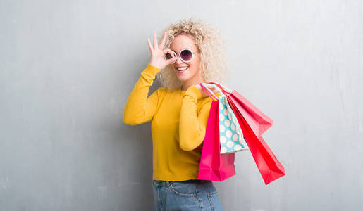 年轻的金发女人，在灰色的背景下，拿着购物袋，销售着，快乐的脸，微笑着，做着好的标志，用手盯着手指。