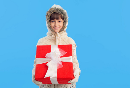 穿着冬天衣服的小女孩手里拿着一个蓝色背景的红色礼品盒。 圣诞礼物。