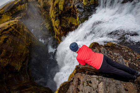一个穿红色衣服的年轻女子正在欣赏躺在岩石上的瀑布的景色。 巨大的水流从岩石中掉下来。 冰岛欧洲