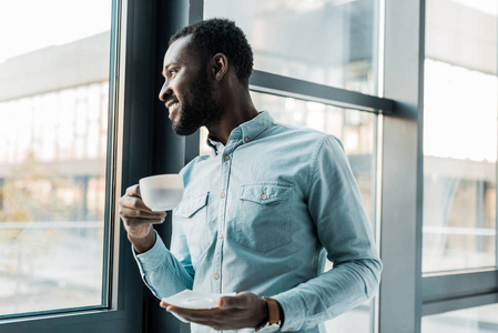微笑的非裔美国人拿着一杯咖啡，望着窗外