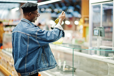 时尚休闲的非裔美国男人，穿着牛仔裤夹克和黑色贝雷帽，拿着篮子，在超市买酒。