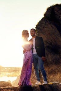 一对相爱的夫妇站在飞溅的水中对抗日落。 一个男人拥抱一个穿着漂亮衣服的女人。 眼睛里的爱