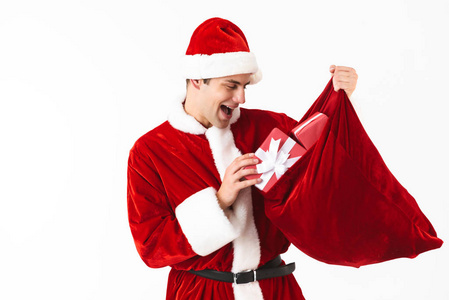 乐观男人30岁的肖像，穿着圣诞老人服装，戴着红色帽子，手里拿着节日袋，礼品盒隔在演播室的白色背景上