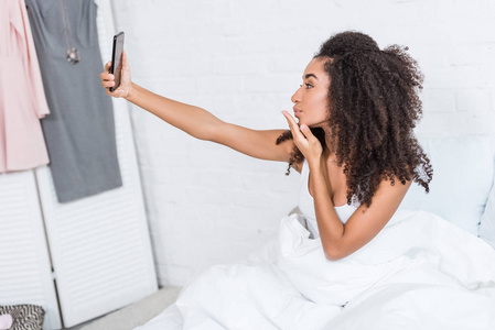 非洲裔美国女孩在家的早晨在床上吹空气吻和自拍智能手机的侧视图