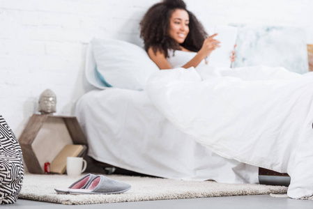 地板上拖鞋的选择性聚焦和非洲裔美国妇女在家后面的床上使用数字平板电脑。