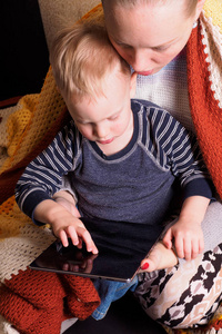 年轻母亲和她的小儿子使用电脑平板电脑的肖像。 幸福的家庭在家里用电子平板电脑一起玩。