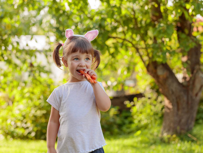 快乐的小女孩吃新鲜甜胡萝卜。 在花园里阳光明媚的夏天，带着兔子耳朵的欧洲小女孩。