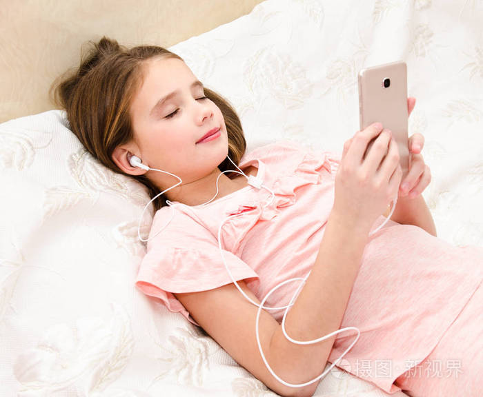 可爱的小女孩躺在床上,用智能手机听音乐,听耳机耳机