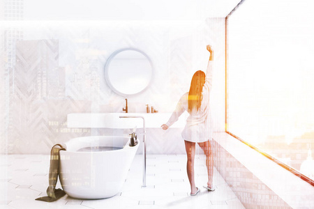 女性在现代浴室的内部与白色木制墙壁瓷砖地板全景窗口，白色浴缸和白色水槽与圆镜以上。 色调图像双曝光