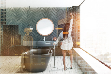 女性在现代浴室的内部与黑色木制墙壁，木制地板，全景窗口，灰色浴缸和灰色水槽与圆形镜子以上。 色调图像双曝光