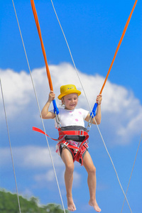 一个小女孩在蹦床上跳得很高，蹦床上用橡胶绳抵着蓝天和白云。 冒险和极限运动。 暑假跳跃的概念。